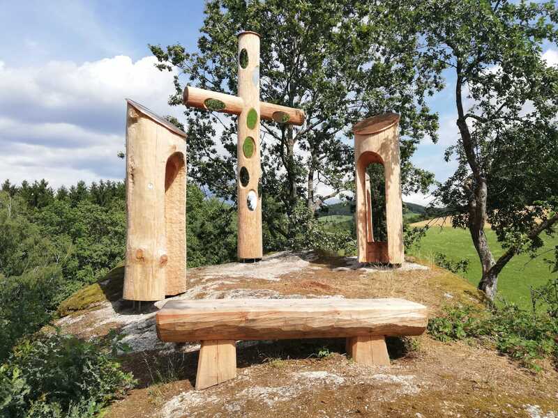 Auf einer Steinformation stehen ein großes Holzkreuz, 2 Holzstelen und eine Holzbank.